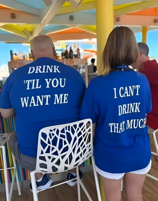 Personer i blå t-shirts med humoristisk text bakifrån, sittande på en bar med färgglada parasoller i bakgrunden.