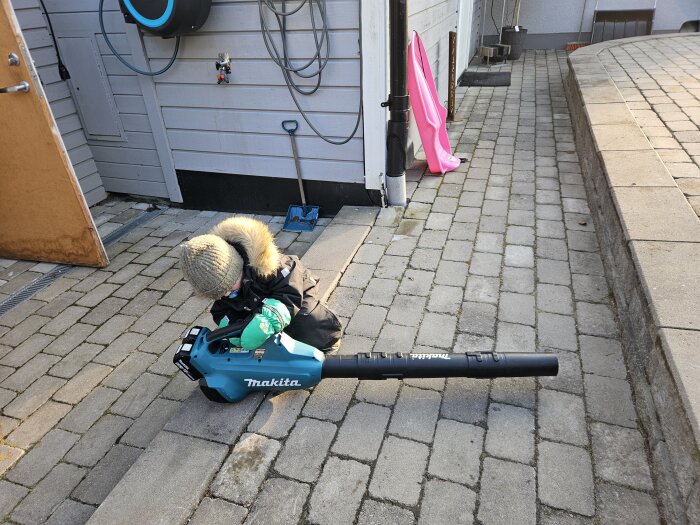 Barn i vinterkläder lekande med en lövblåsare på stenläggning vid husvägg.