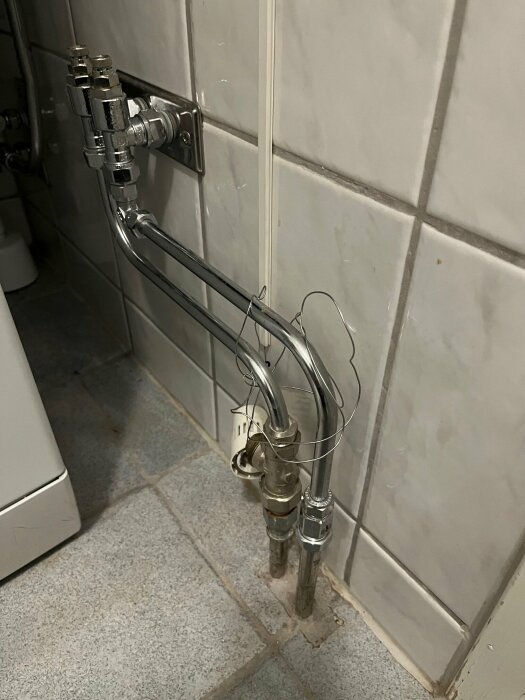 Rör och anslutningar för golvvärmesystem på badrumsvägg, med termostat som verkar löst hängande. Kakelvägg och klinkergolv syns.