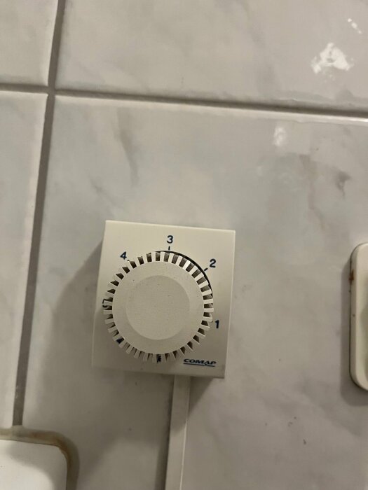 Termostat med ratt monterad på kaklad vägg, inställningarna går från 1 till 4, märkt "COMAP".