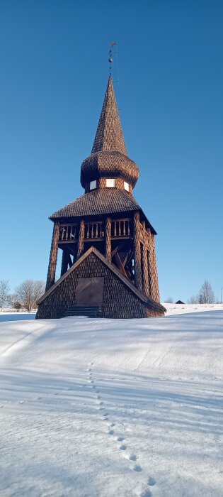 Träpavillon med spetsigt torn på en snötäckt kulle under klarblå himmel, fotspår leder fram till strukturen.