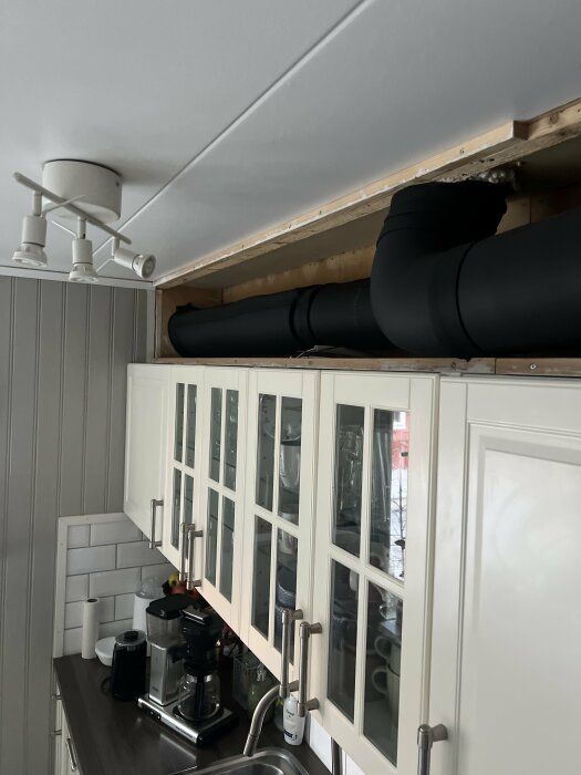 Svart ventilationsrör installerat ovanför köksskåp i kök med vit inredning. Spotlights i taket ovanför.