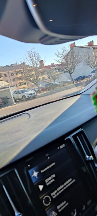 Vy inifrån en bil, parkering med en Volkswagen och flera parkerade bilar framför ett flerfamiljshus syns genom framrutan.