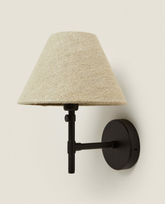 Vägglampa med beige textilskärm och svart arm, skapar en mysig känsla i rummet.