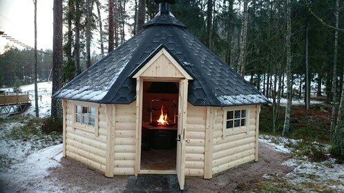 Stuga av trä med sexkantigt tak, belägen i skogsmiljö med snö på marken. Inne i stugan syns en eldplats i mitten.