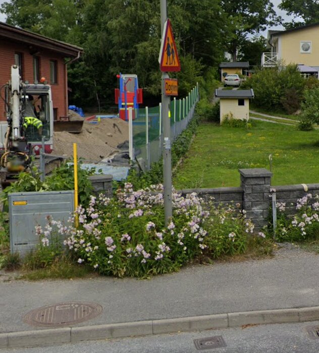 Gatan med ett vägarbete, blommande buskar och en stolpe med skylt. Ett staket och en gräsmatta med ett litet hus i bakgrunden.