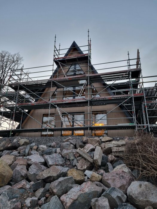 En triangelformad träbyggnad med ställningar runt, med svarta vindskivor och takpannor på plats mot en molnig himmel.