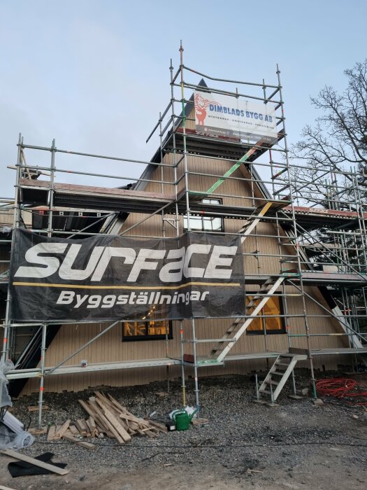 Byggarbetsplats med ställningar uppställda runt ett hus under renovering, svarta vindskiveplåtar monterade, brädhög framför byggnaden.