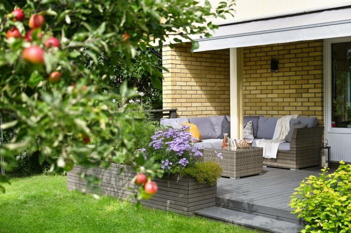 Uteplats med rottingmöbler under en utbyggd balkong, två synliga stolpar och blommor i fokus.