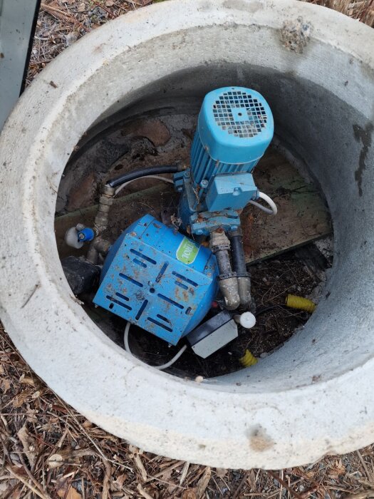 Gamla pumpmotor och kopplade rör i en borrad brunn, omgiven av naturmaterial och jord.
