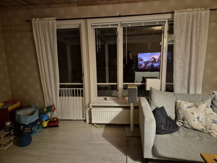 Vardagsrum med grå soffa, fönster med vita gardiner, leksaker på golvet och en element under fönstret. Reflektion av TV på fönstret.