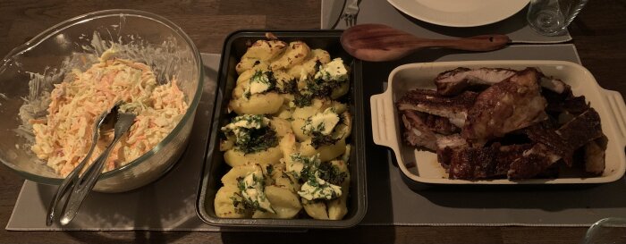 Coleslaw i skål, ugnsrostad potatis med persiljesmör, och spareribs i ugnsform på ett matbord.