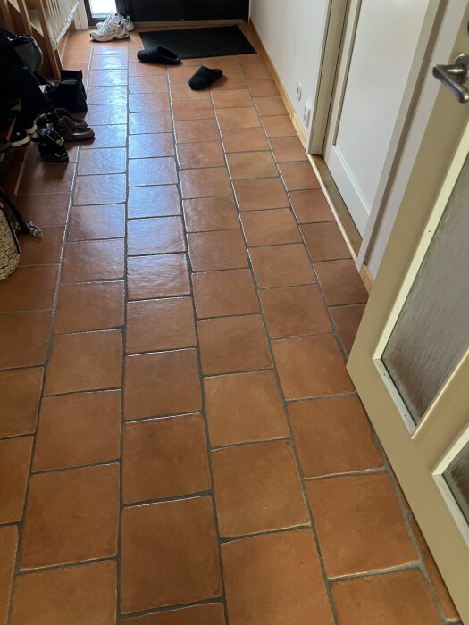 Gul/orange kakelgolv i hallen framför en dörr, med några skor och tofflor på golvet.