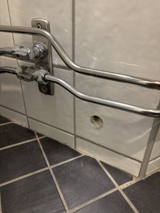 Hål i kaklad vägg nära golvet i ett badrum med två metallrör ovanför, som har fyllts delvis med grå silikon.