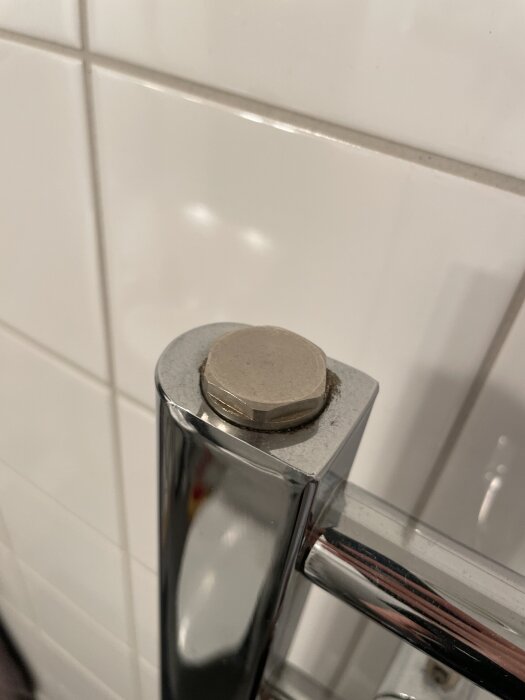 Kromad handdukstork med en stor skruvmutter upptill som saknar ett täcklock, mot en kaklad vägg i bakgrunden.