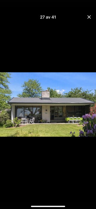 Villa med stora glaspartier, vit fasad, trädgård med utemöbler och grön gräsmatta under klarblå himmel.