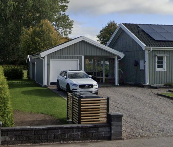 Grått garage med carport och vit bil parkerad framför; byggnad bredvid garaget, gräsmatta och grusad uppfart.