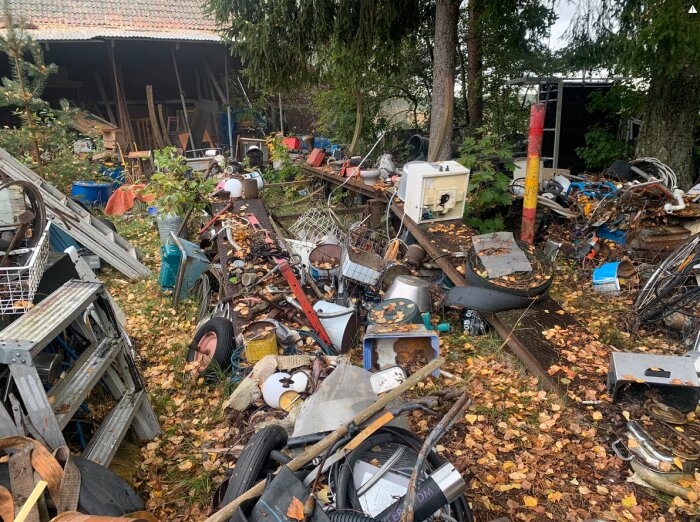 Övergiven tomt med bråte och skrot bakom ett hus med trasigt tak, täckt av höstlöv och omgiven av träd och växtlighet.