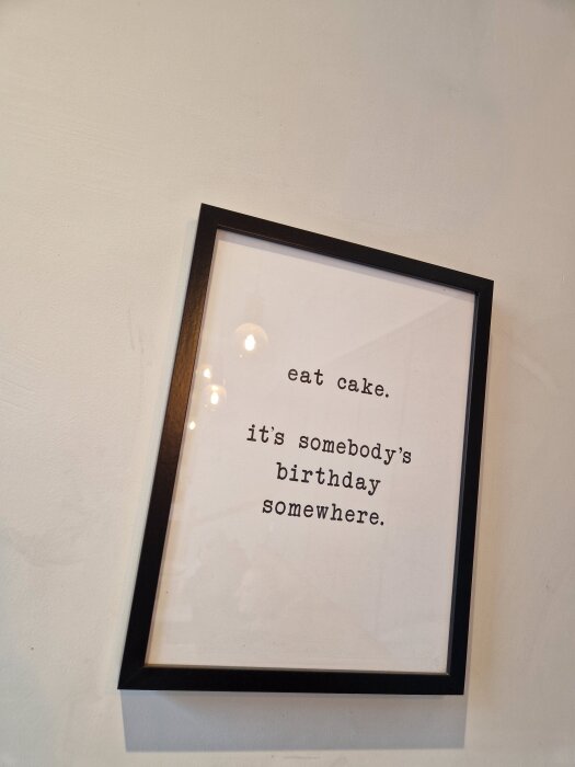 Tavla på väggen med texten "eat cake. it's somebody's birthday somewhere." mot en vit bakgrund.