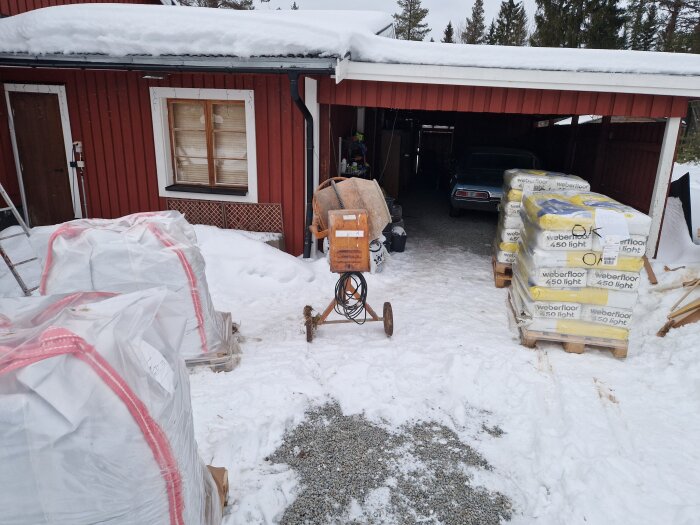 Snötäckt gårdsplan med byggmaterial, flera pallar Weber golvavjämning och storsäckar, samt en cementblandare framför ett rött hus.