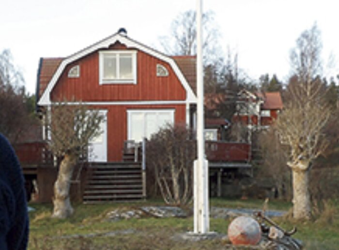 Ett rött trähus med vita knutar i förgrunden, omgivet av träd och buskar, med en vit flaggstång framför. Ett annat hus syns i bakgrunden.