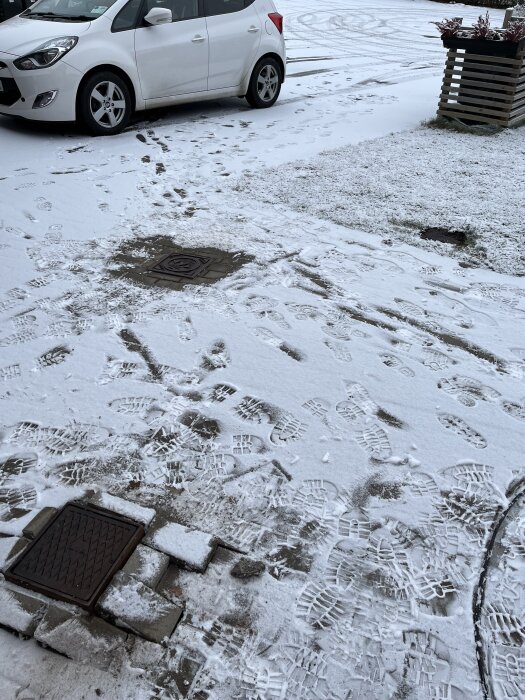 Snötäckt mark med två brunnslock, varav ett upphöjt, fotspår och en parkerad vit bil i bakgrunden.