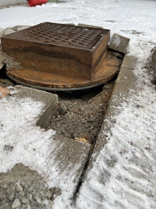 Brunnlock i metall omgiven av is, något upphöjt över stensättning, med snö på marken runt omkring.