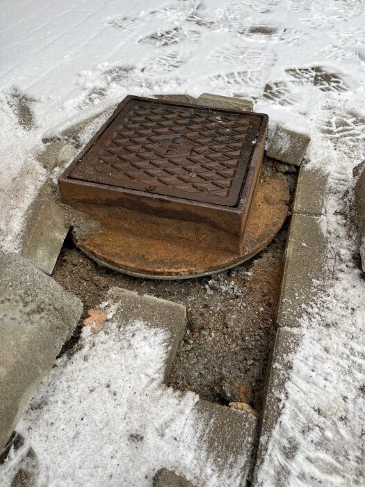 Fyrkantigt metallbrunnslock inbäddat i stenlagd yta, delvis täckt av is och snö, med synlig höjdskillnad.