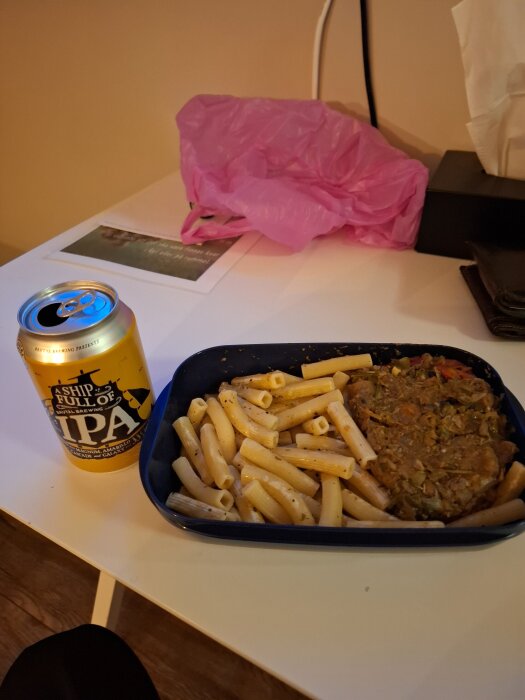 Matlåda med pasta och ragu bredvid en burk IPA på ett bord. I bakgrunden ligger en rosa plastpåse och några papper.