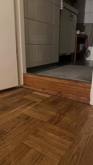 Ingång till badrum med tröskel som är cirka 15 cm högre än angränsande trägolv, synliga kakelväggar och badrumsmatta i bakgrunden.