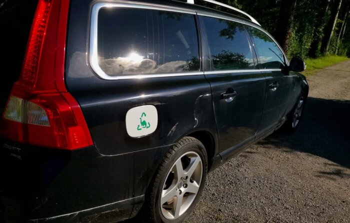 Svart Volvo V70 parkerad utomhus, solreflektion i sidofönstret, skog i bakgrunden, synlig bensinlucka med symbol.