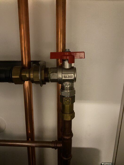 Avstängningsventil på kopparrör med rött handtag, svårtillgänglig på grund av närliggande rör, i ett köksutrymme.