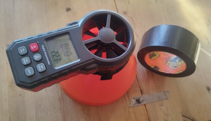 Digital anemometer på en orange kon bredvid en rulle svart tejp på ett träbord.