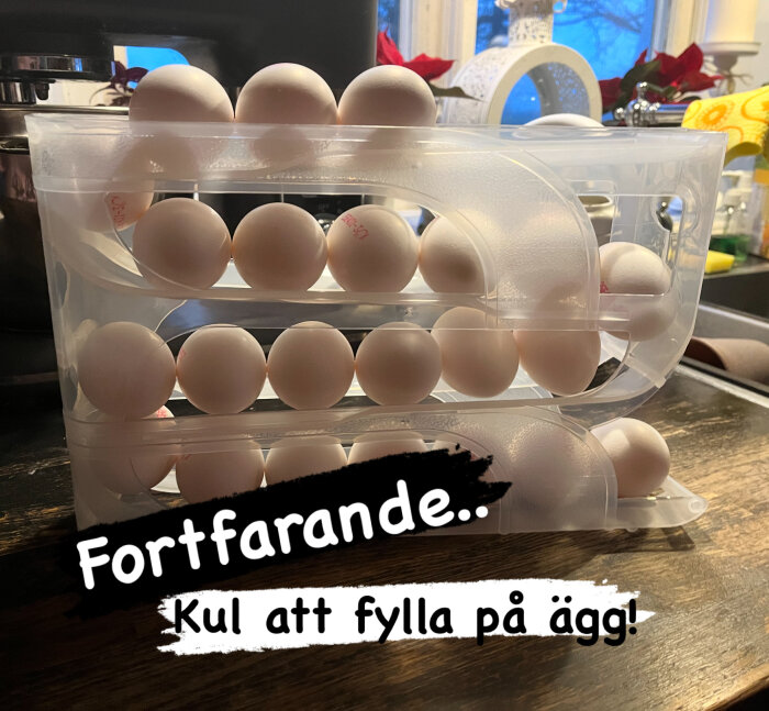 Ägg förvaras i en genomskinlig plastlåda på ett köksbord med texten "Fortfarande.. kul att fylla på ägg!" i förgrunden.