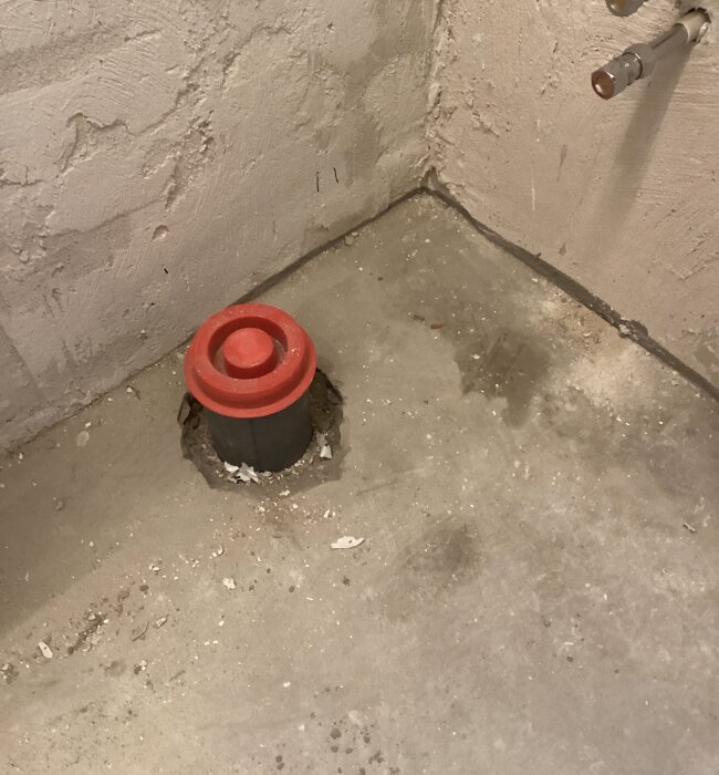 Avloppsrör med röd propp i ett renoverat badrumsgolv, synlig muff under flytspacklad yta nära väggen.