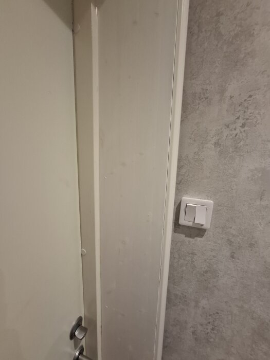 Närbild på en dörrpost med gula fläckar under vit färg, vid sidan av en ljusgrå vägg och en strömbrytare.