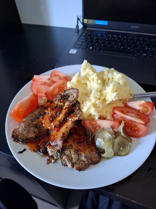 Tallrik med grillat kött, gräddig potatismos, skivade tomater och picklad gurka på ett bord bredvid en bärbar dator.