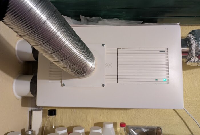 Installationskit för ventilation med ljuddämpare, en stor spiralrörsanslutning syns monterad på en vit enhet på en vägg i ett förråd.