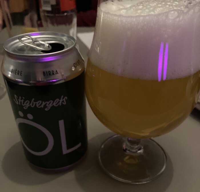 En ölburk med texten "Stigbergets ÖL" bredvid ett glas fyllt med skummande öl, placerat på ett bord.