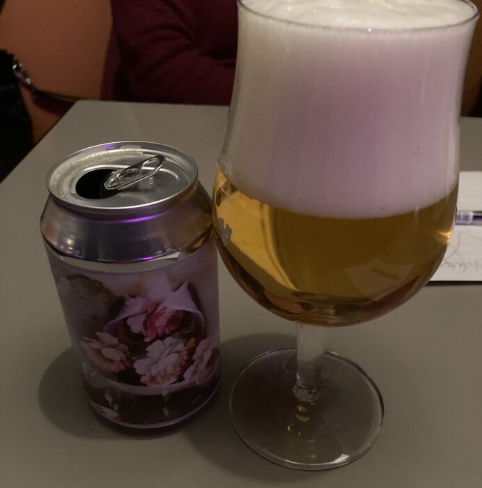 Öl på burk med blomsterdesign bredvid ett glas fyllt med skummande öl på ett bord.