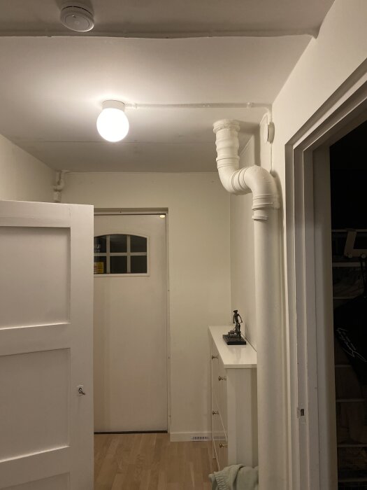 Vitt avloppsrör längs taket och väggen i ett badrum med vitt kakel och en sak på en byrå.