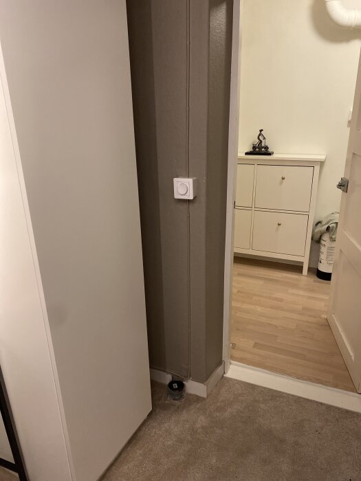 Gammalt avloppsrör vid golvet mellan två rum, synligt i hörnet under ett grått vägguttag i en hall med parkettgolv och beige matta.