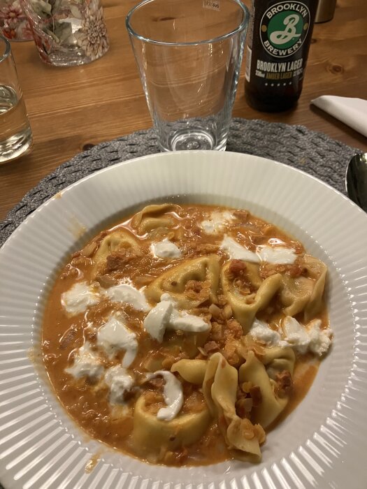 Soppa med pastakuddar, hackad chorizo, lök, vitlök, krossade tomater, grädde och riven mozzarella i en vit skål. Flasköl och glas i bakgrunden.