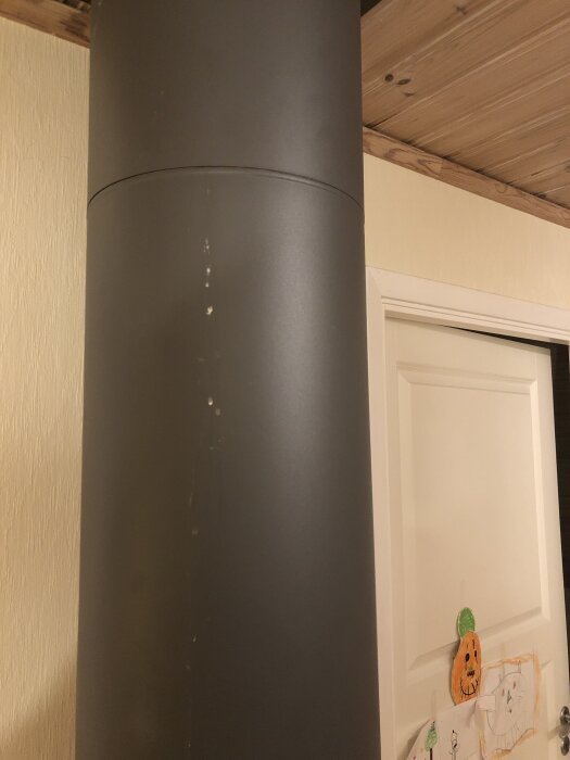 Svart rör mot träpaneltak med vita fläckar vid skarven, ovanför en dörr med barnteckningar.