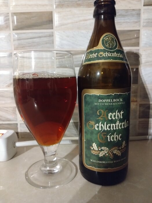 En flaska och glas med Aecht Schlenkerla Eiche Doppelbock öl på ett köksbänk med kaklad bakgrund.