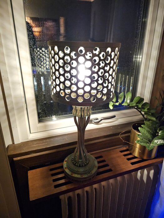 Egendesignad lampa med perforerad lampskärm i PETG, placerad på fönsterbräda bredvid en grön växt i en metallkruka.