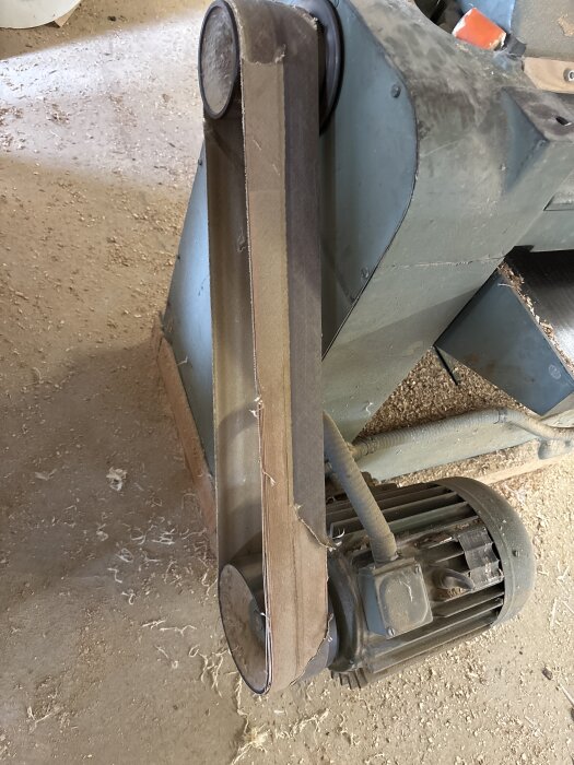 En sliten slipmaskin med sandpapper och motor, omgiven av träspån på ett verkstadsgolv.
