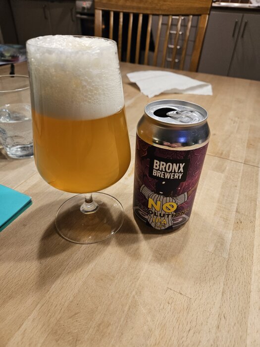 Ett glas med skummande öl bredvid en öppnad burk "Bronx Brewery No Resolutions IPA" på ett träbord.