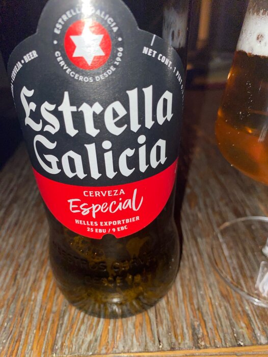 Flaska Estrella Galicia Cerveza Especial bredvid ett ölglas på träbord.