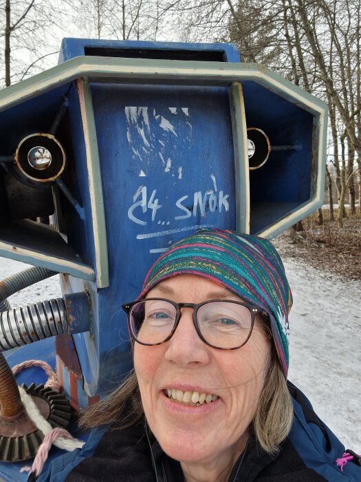 Person framför en stor, blå skulptur med text på och träd i bakgrunden, på en vinterdag med snötäckt mark.